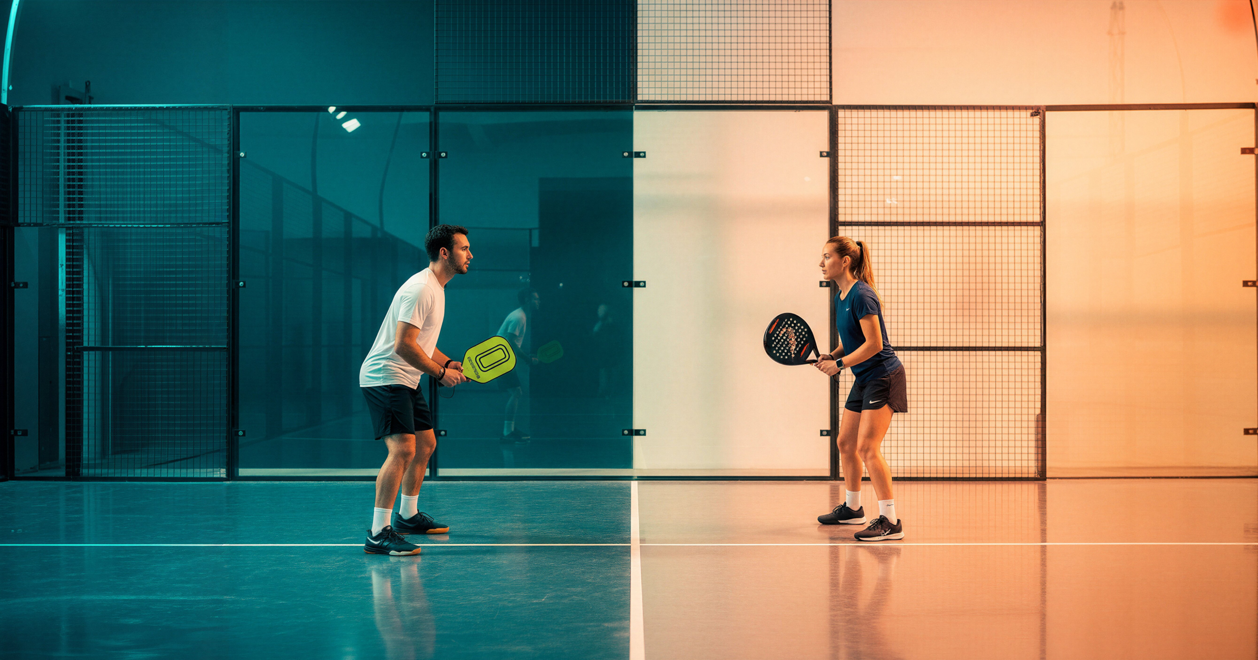 A man and woman face each other on a court for padel vs pickleball, divided by striking teal and orange lighting.