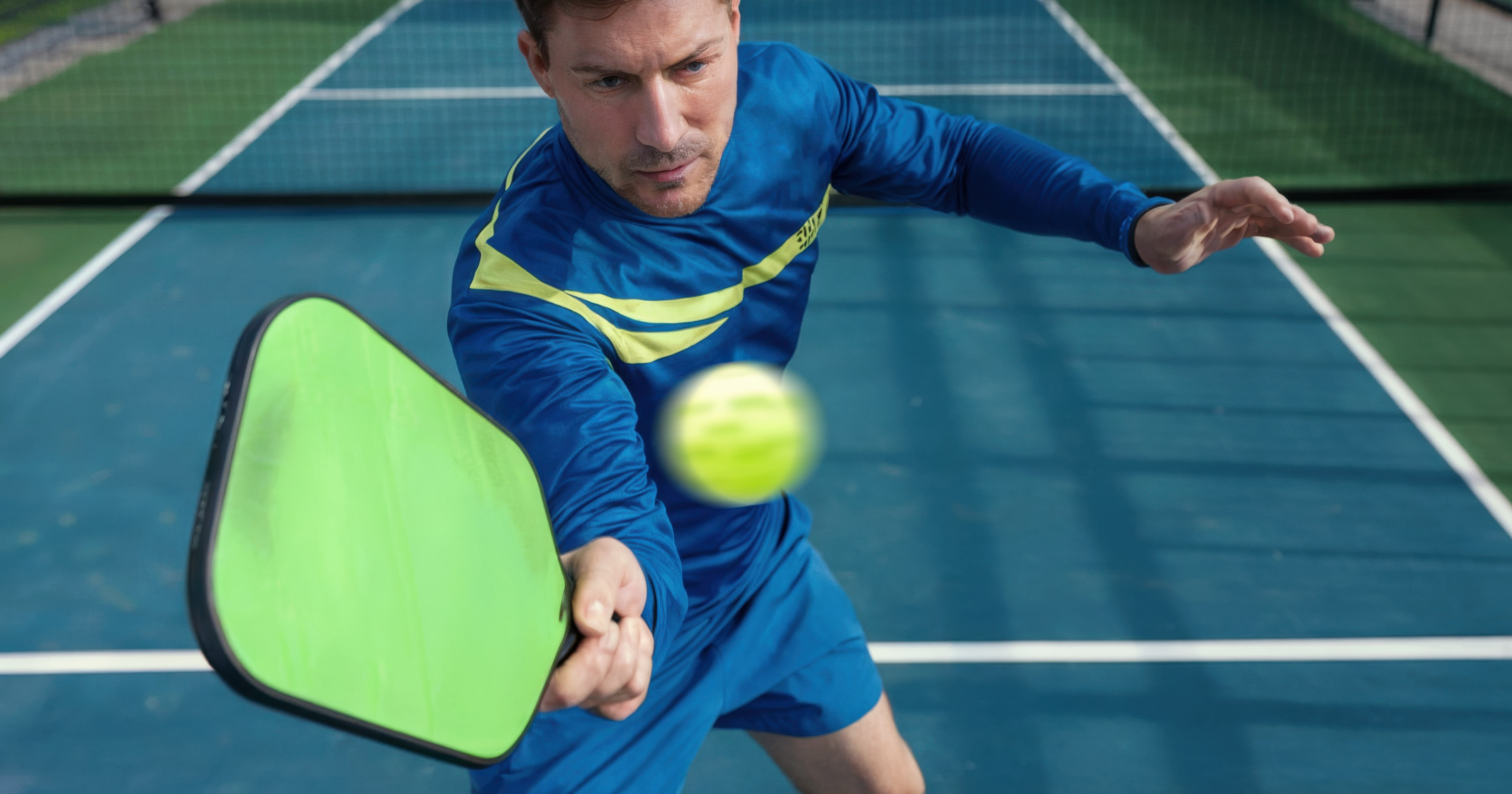A player in blue swings a green paddle at a ball. Many wonder why is it called pickleball while watching the fast action.