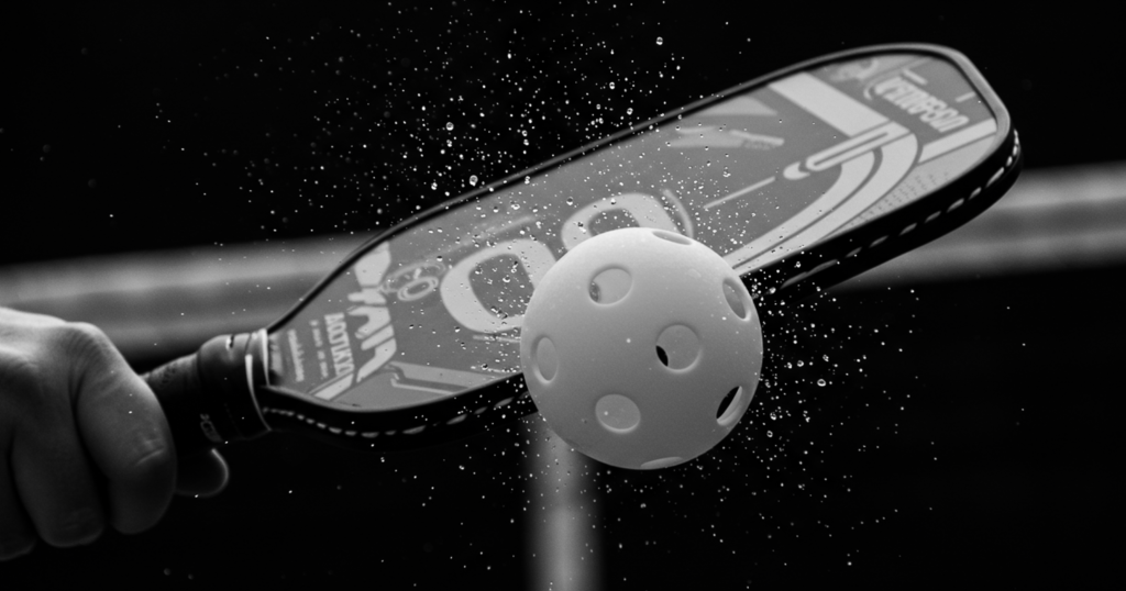Black and white close-up of a paddle hitting a pickleball, creating a dramatic spray of water droplets.