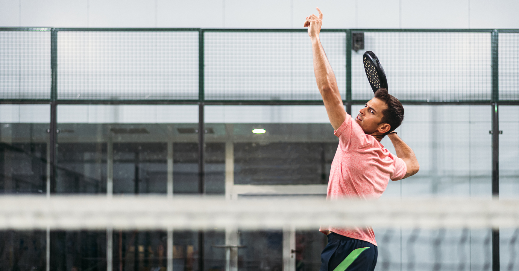 Male pickleball player performing an overhead serve on an indoor court with a paddle raised behind his head.