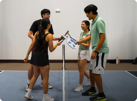 Four players smiling and tapping their paddles over the net following a game on an indoor pickleball court.
