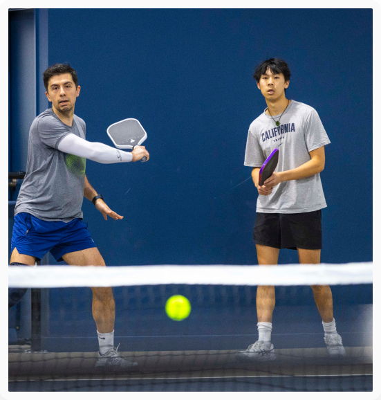 Two men playing a doubles pickleball match indoors, with one player swinging his paddle while his teammate watches the ball cross the net.