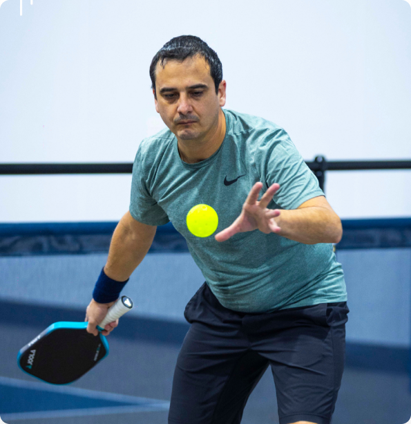 A man in a green shirt tracked mid-motion as he focuses closely on a bright yellow pickleball flying toward him.