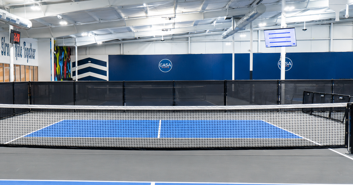 Indoor pickleball court with a black net and blue playing area.