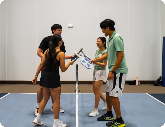 Four players smiling and tapping their paddles over the net following a game on an indoor pickleball court.