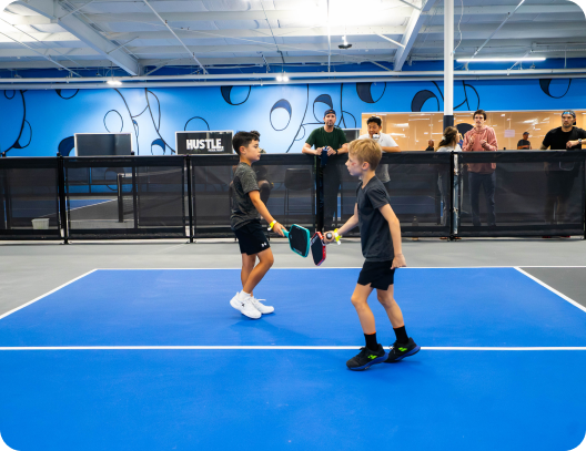Two young boys tap their paddles together over the net on a bright blue indoor pickleball court with spectators looking on from behind a barrier.