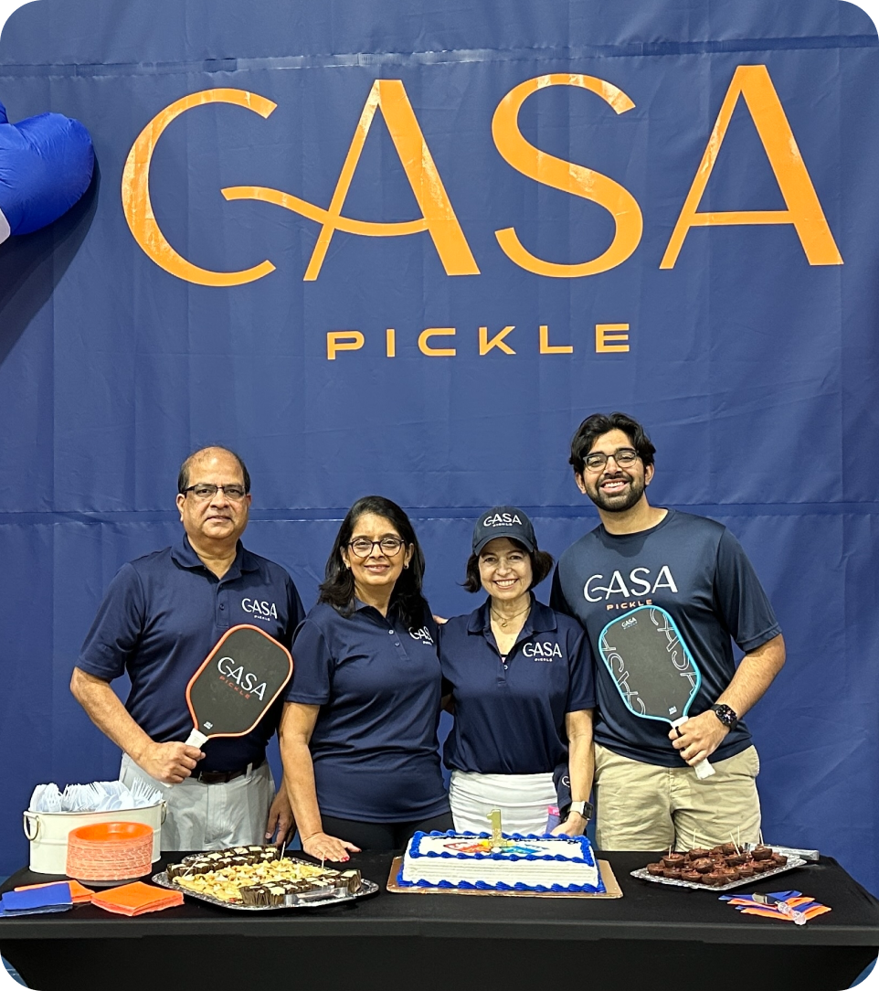 A group of four people posing behind a table with a cake and snacks in front of a blue "CASA PICKLE" backdrop.
