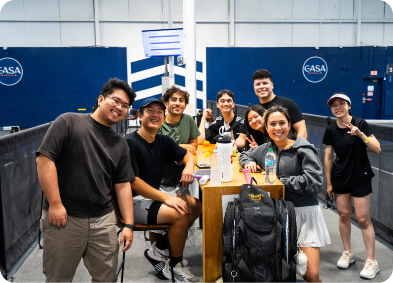 A diverse group of young adults smiling and posing around a high wooden table next to an indoor pickleball court.