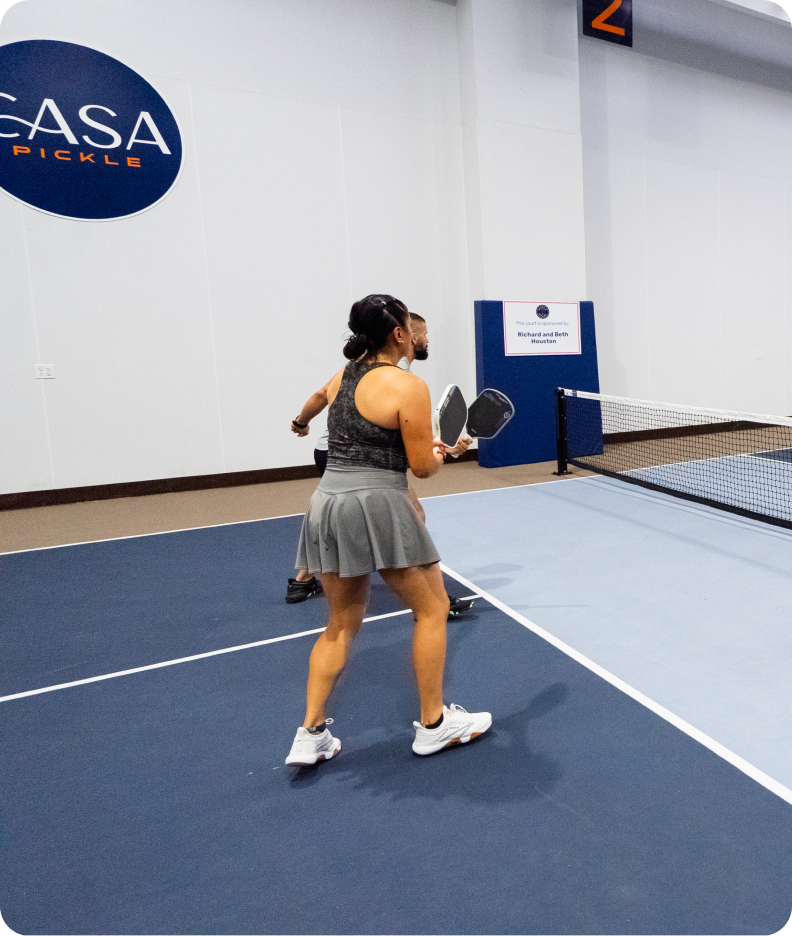 A woman in a grey athletic skirt and tank top prepares to hit a pickleball during a doubles match on an indoor court.