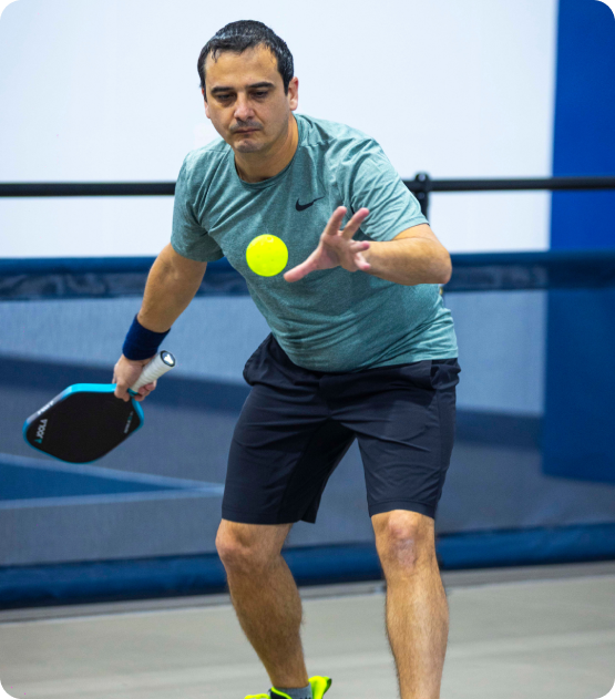 A man in a green shirt tracked mid-motion as he focuses closely on a bright yellow pickleball flying toward him.