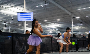 Two women in athletic skirts ready their paddles to receive a pickleball on an indoor court.