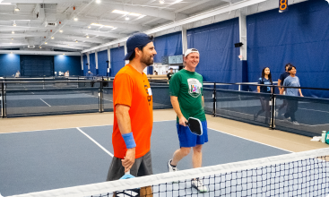Two men wearing t-shirts and caps smile and talk while walking off an indoor pickleball court.
