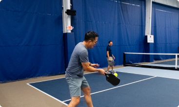 A side-view shot of a man in a grey shirt preparing to strike a pickleball on an indoor court.