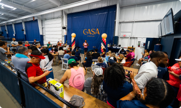 A large crowd sits in a spectator area watching a presentation in front of a blue "CASA PICKLE" wall banner.