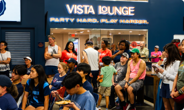A crowded indoor social gathering with people sitting and standing beneath a glowing sign that reads "VISTA LOUNGE PARTY HARD. PLAY HARDER."