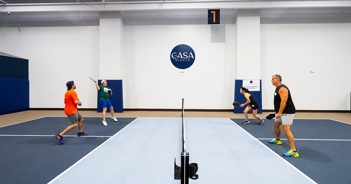 Four adults playing doubles pickleball on an indoor court.