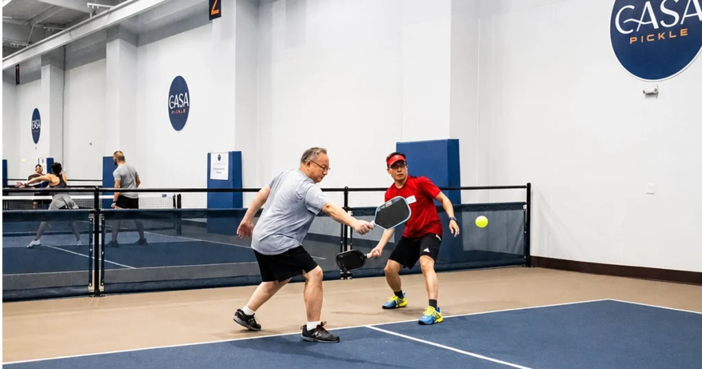Two men practice pickleball on an indoor court, with a yellow ball midair and other players in the background.