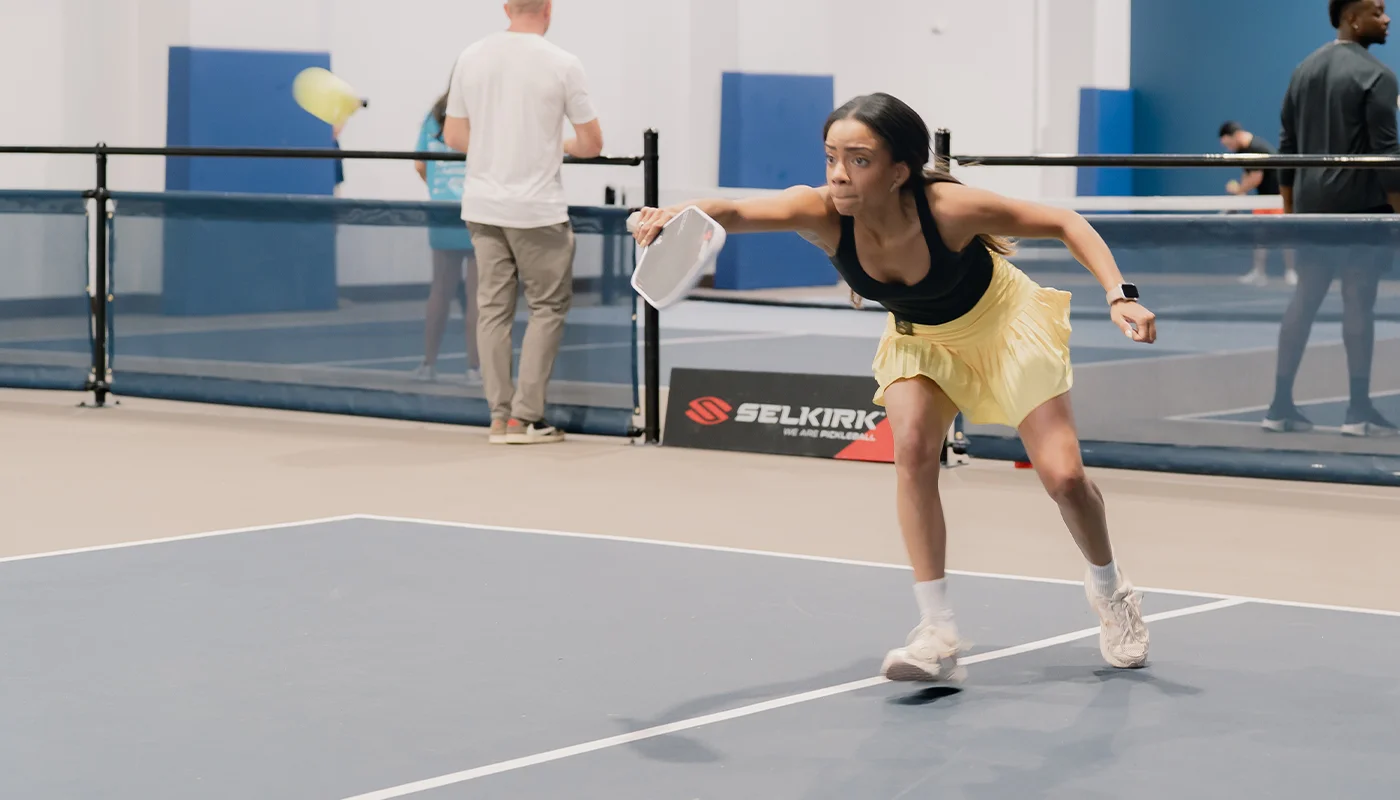 Woman in yellow skirt reaching forward playing indoor pickleball game.