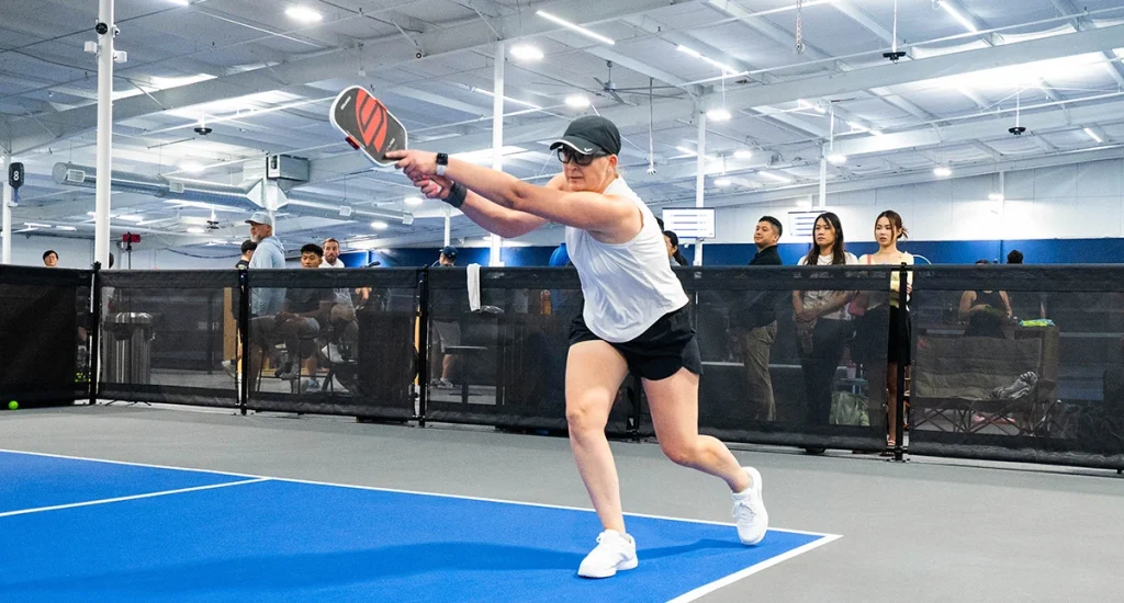 Woman stretching to reach a backhand shot during an indoor pickleball game.
