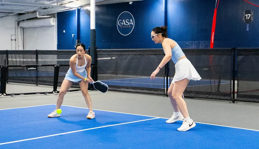 Two women playing a doubles pickleball match on an indoor blue court.